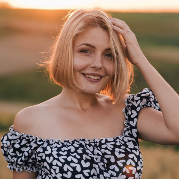 A rejuvenated blond woman in a floral dress, treated by Dr. Jack Peterson's Plastic Surgery clinic, stands in a field at sunset after Restylane® Lyft treatment.