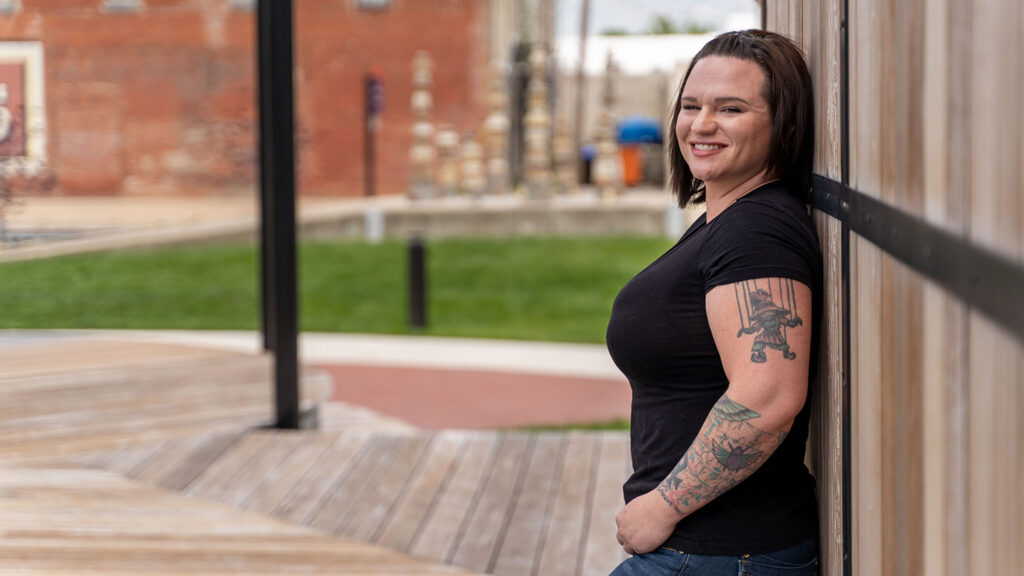 Person with tattoos on both arms leans against a wooden wall outdoors, smiling. Brick and grass background with a blurred figure in the distance.