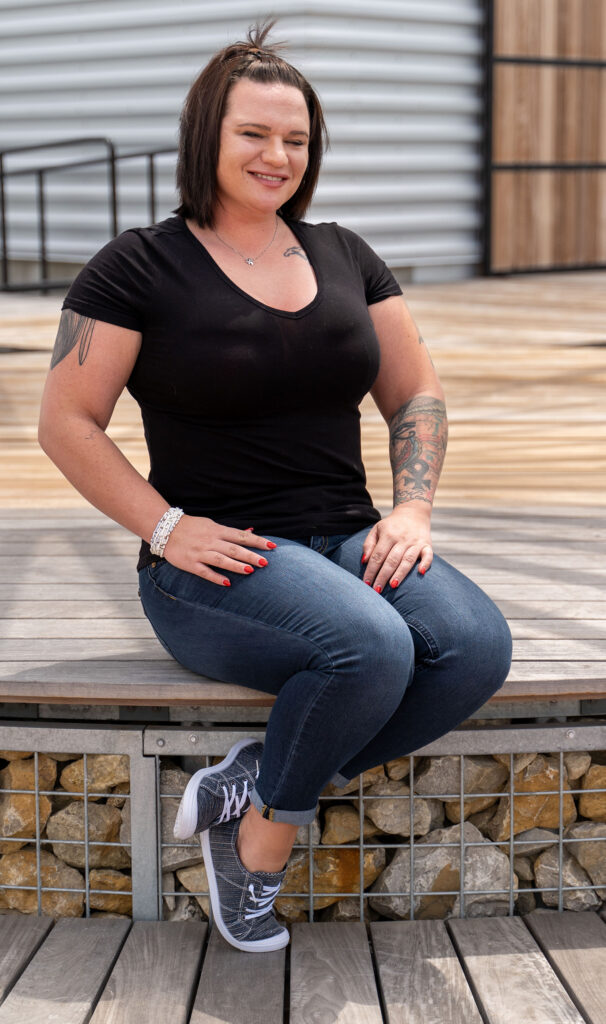 A person with tattoos, wearing a black t-shirt, jeans, and sneakers, sits on a wooden bench outdoors, smiling at the camera.