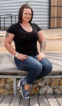 A person with tattoos, wearing a black t-shirt, jeans, and sneakers, sits on a wooden bench outdoors, smiling at the camera.