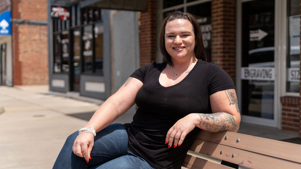Person wearing a black t-shirt and jeans sits on a bench. They have tattoos on their arm and are in front of brick buildings.