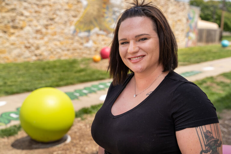 Person with short brown hair and a tattoo on their arm, smiling outdoors in front of a stone wall and colorful playground balls.