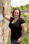 A person in a black t-shirt stands outdoors, leaning against a textured wall, smiling at the camera. The background shows a grassy area with colorful spherical objects.