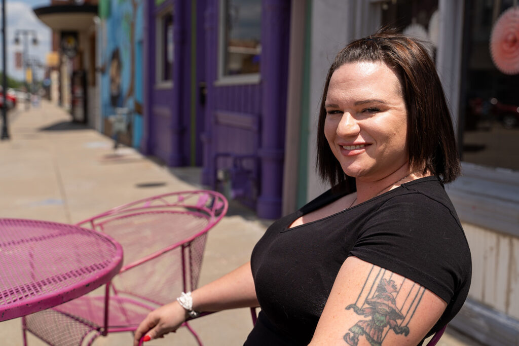 A person with a tattoo on their arm is sitting at a pink outdoor table on a sunny day, in front of a building with vibrant purple and blue walls.