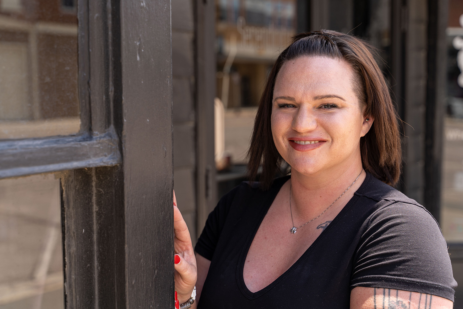 A person with brown hair in a black shirt smiles while standing beside a dark-colored window frame, with a street visible in the background.