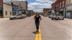 A person stands smiling on a yellow line in the middle of a wide street lined with parked cars and buildings on a clear day.