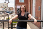 Woman with tattoos and black T-shirt stands outside, leaning on a railing, smiling at the camera. Buildings and parked cars are visible in the background.