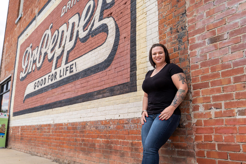 Person in a black t-shirt and jeans stands against a brick wall with a vintage Dr Pepper sign.