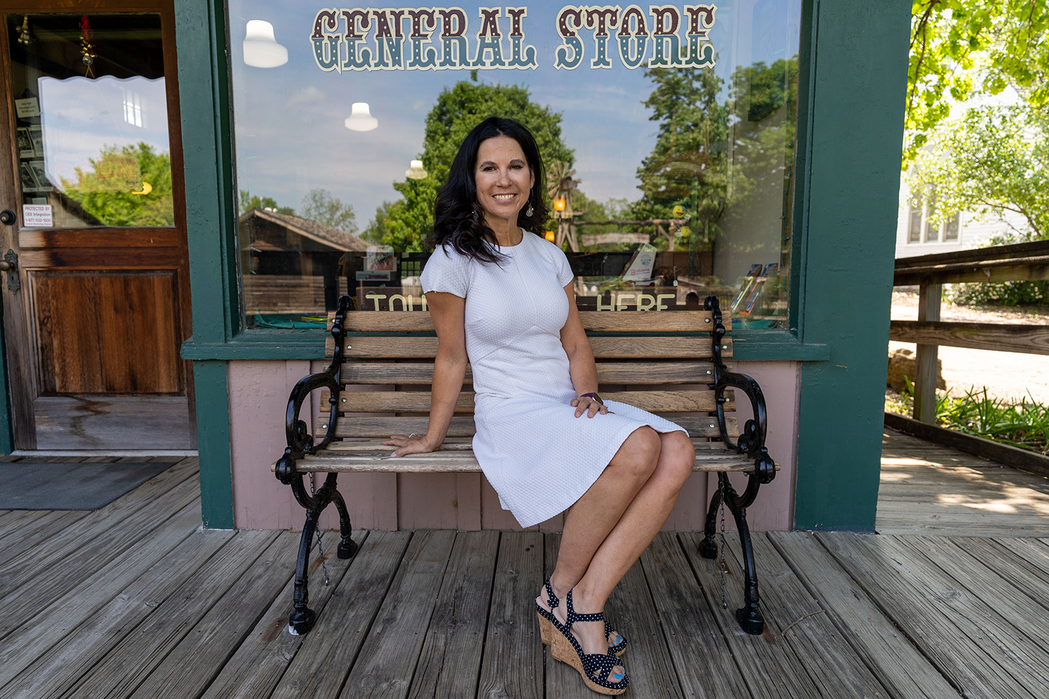 A woman in a white dress and sandals sits on a bench outside a general store.