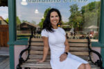 A woman in a white dress sits on a wooden bench in front of a store. There is greenery and buildings reflected in the window behind her.