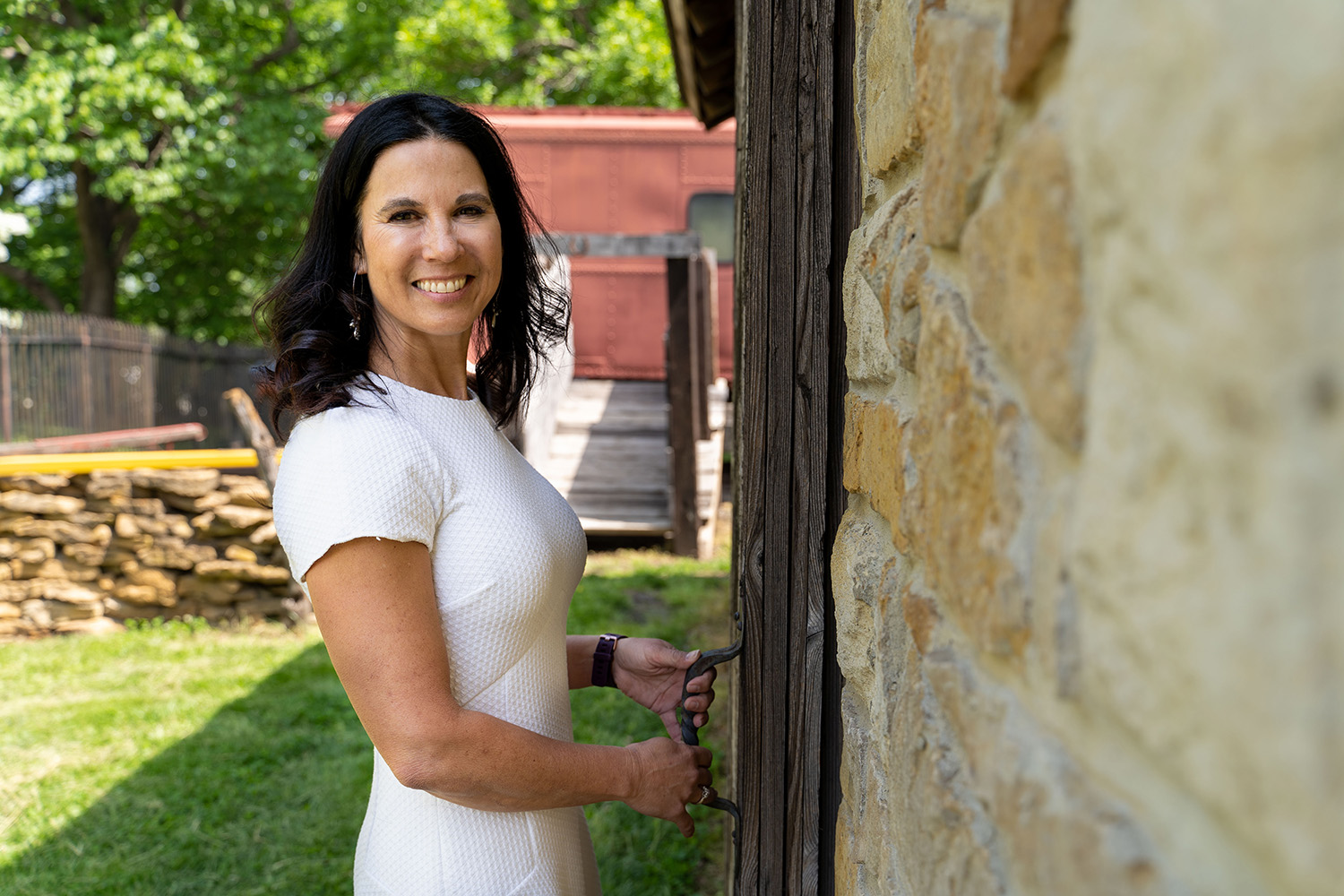 A woman in a white dress smiles while standing next to a wooden door, with a stone wall and greenery in the background.