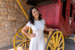 A woman in a white dress stands smiling beside a red stagecoach with yellow wheels against a stone wall backdrop.