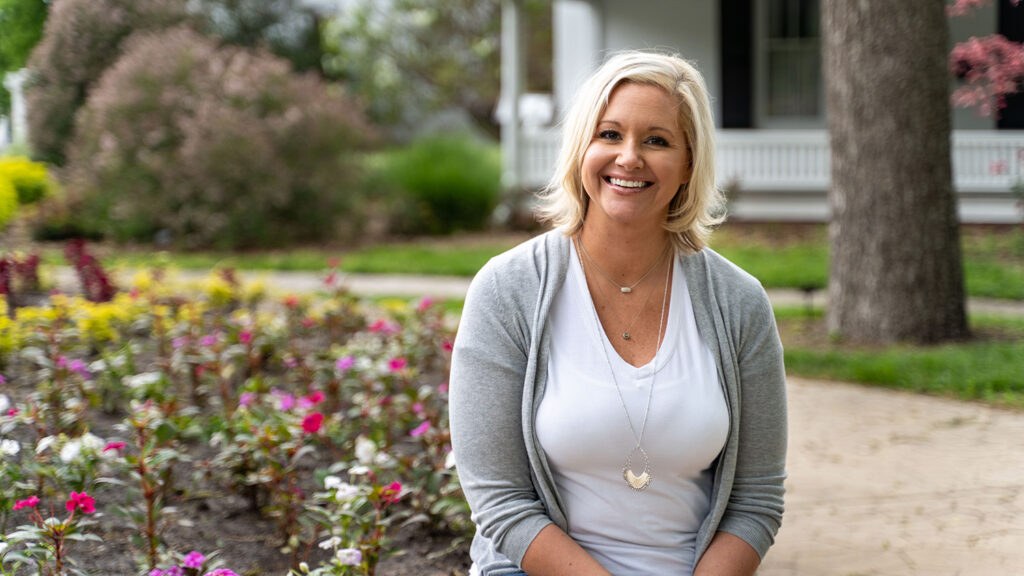 Woman sitting on a park bench, smiling, with colorful flowers in the foreground and a house in the background.
