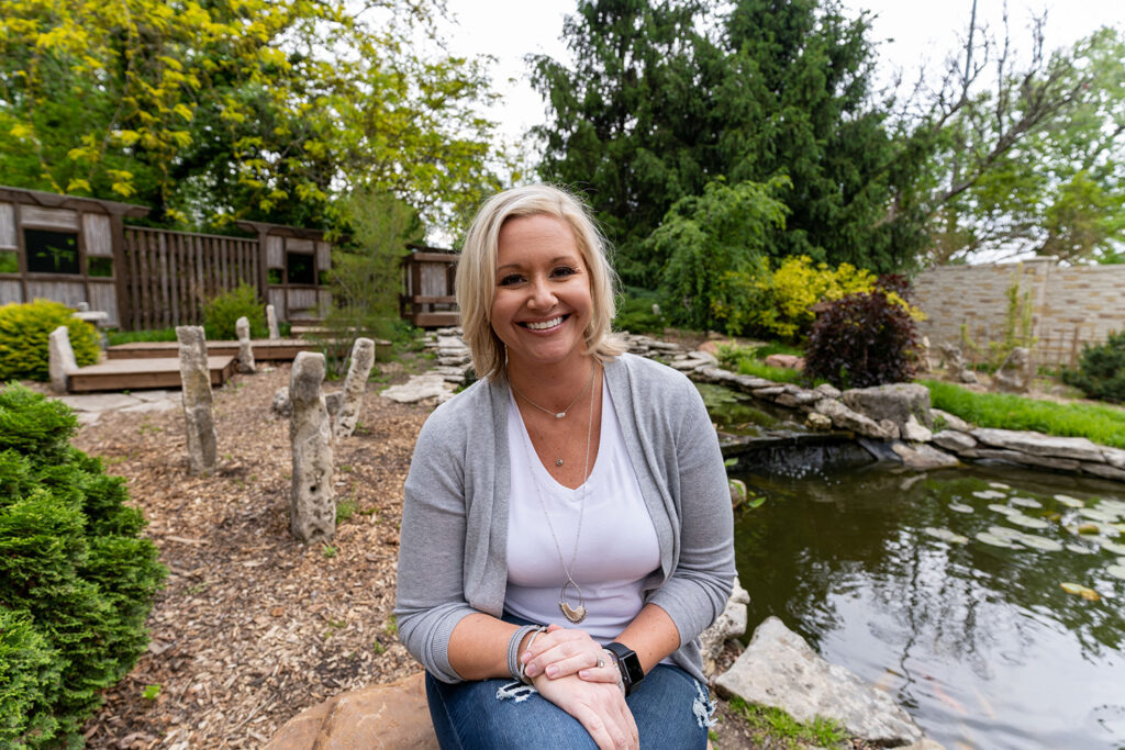 A woman with blonde hair sits smiling by a pond in a garden with trees, rocks, and wooden structures in the background.