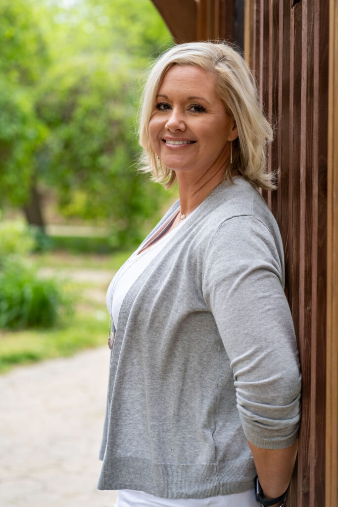 Smiling person with short blond hair leans against a wooden wall outdoors, wearing a gray cardigan over a white top.