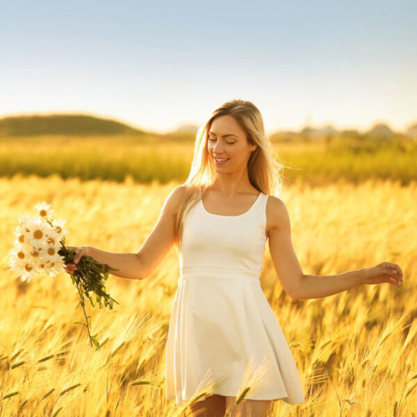 A woman in a white dress stands confidently in a golden wheat field, holding daisies, basking in sunlight, showcasing her serene confidence after choosing Breast Implant Removal.