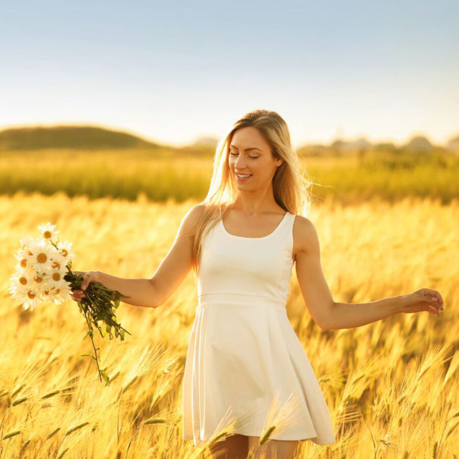 A woman in a white dress stands confidently in a golden wheat field, holding daisies, basking in sunlight, showcasing her serene confidence after choosing Breast Implant Removal.