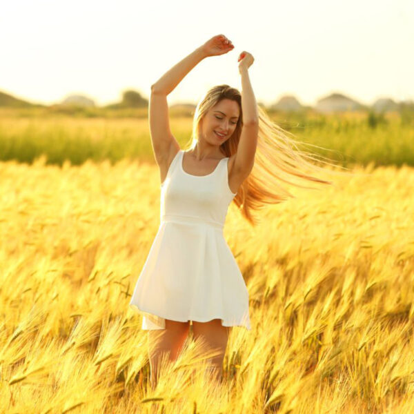 A woman stands confidently in a golden wheat field with arms raised, smiling and enjoying the sunlight after undergoing Breast Implant Removal.