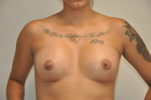 A woman with visible tattoos on her chest and arms stands topless against a plain background.