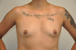 A topless person with chest tattoos and nipple piercings stands facing forward against a plain background.