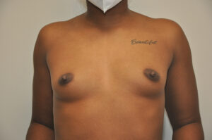 A person with a medium skin tone stands shirtless, wearing a white face mask. The word "Beautiful" is tattooed on their upper right chest.