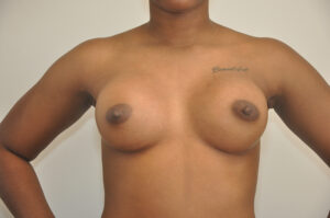 A woman with a "beautiful" tattoo on her shoulder stands topless against a plain background, arms relaxed at her sides.