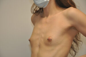 A person with long hair and a mask is shown shirtless from the shoulders up, displaying nipple piercings.