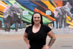 A woman with tattoos stands smiling in front of a mural featuring two painted elephants and colorful abstract shapes, her glowing complexion hinting at the benefits of skin treatments for acne scarring.