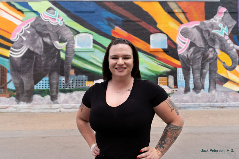 A woman with tattoos stands smiling in front of a mural featuring two painted elephants and colorful abstract shapes, her glowing complexion hinting at the benefits of skin treatments for acne scarring.