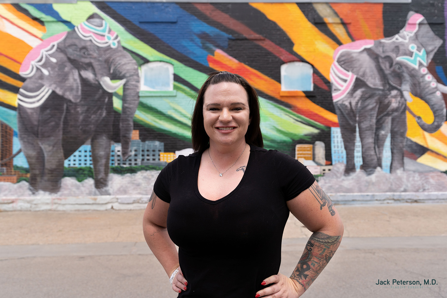 A woman with tattoos stands smiling in front of a mural featuring two painted elephants and colorful abstract shapes, her glowing complexion hinting at the benefits of skin treatments for acne scarring.