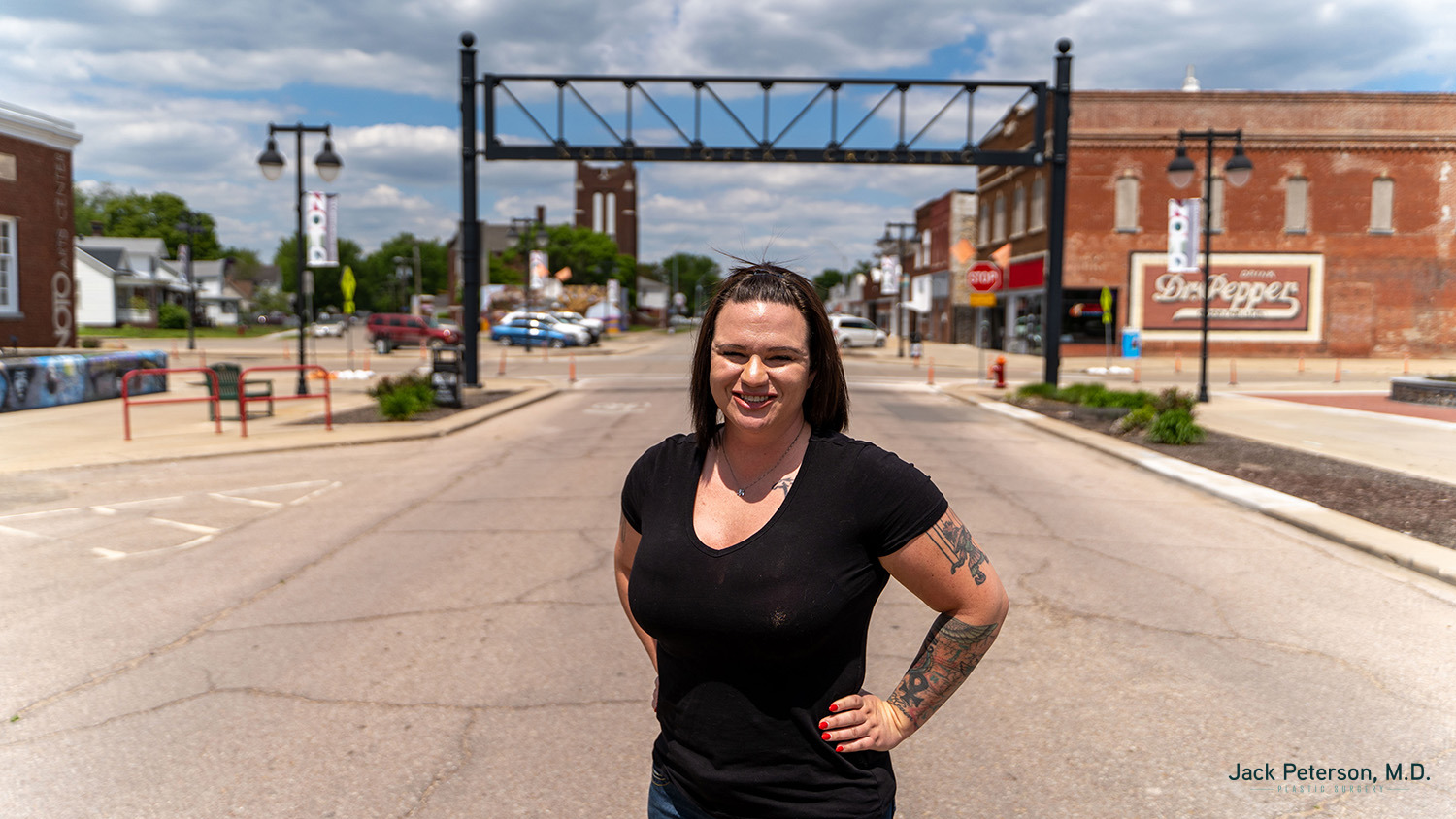 A woman with tattoos stands smiling in the center of a small town street, glowing as if she just perfected her skincare routine after chemical peel, with brick buildings and signs visible in the background on a sunny day.
