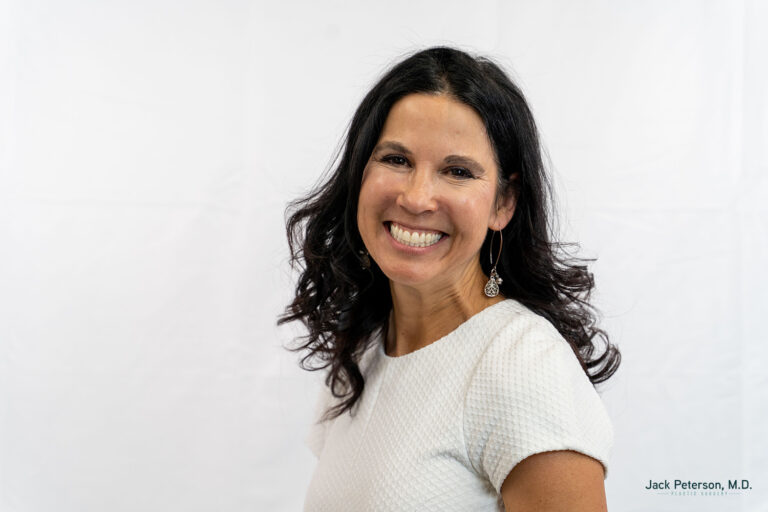Woman with long dark hair wearing a textured white short-sleeve top, smiling, standing against a plain white background—radiating confidence like those who have explored personalized mommy makeover options.
