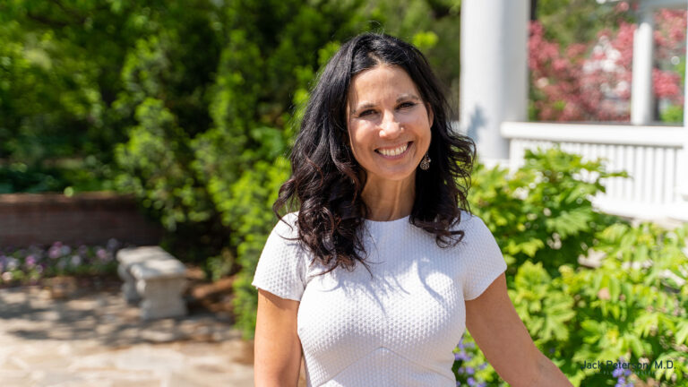 A woman with long dark hair, wearing a short-sleeved white textured dress, smiles while standing outdoors in a sunny garden, her glowing complexion reflecting the best skin treatments for acne.