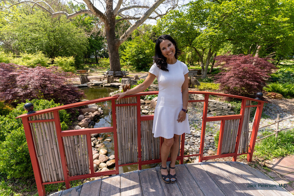 A woman in a white dress stands on a small red wooden bridge in a garden with trees, shrubs, and a pond—a peaceful spot perfect for reflecting on skin treatments for acne scarring.