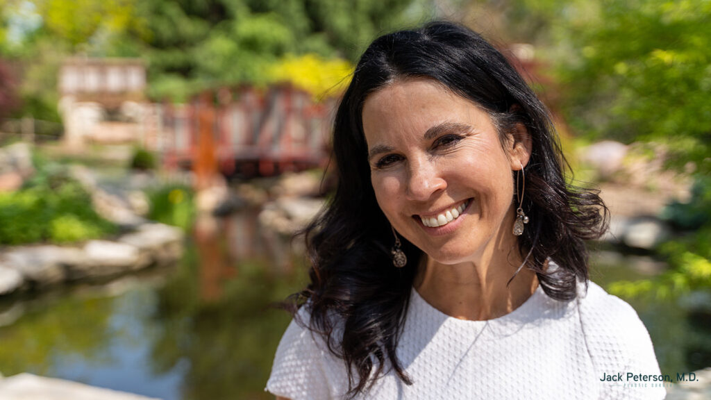 A woman with long dark hair, wearing a white textured top, smiles outdoors near a pond and garden with lush greenery, highlighting the natural beauty achieved through non-surgical facelift options. A blurred red structure appears in the background.
