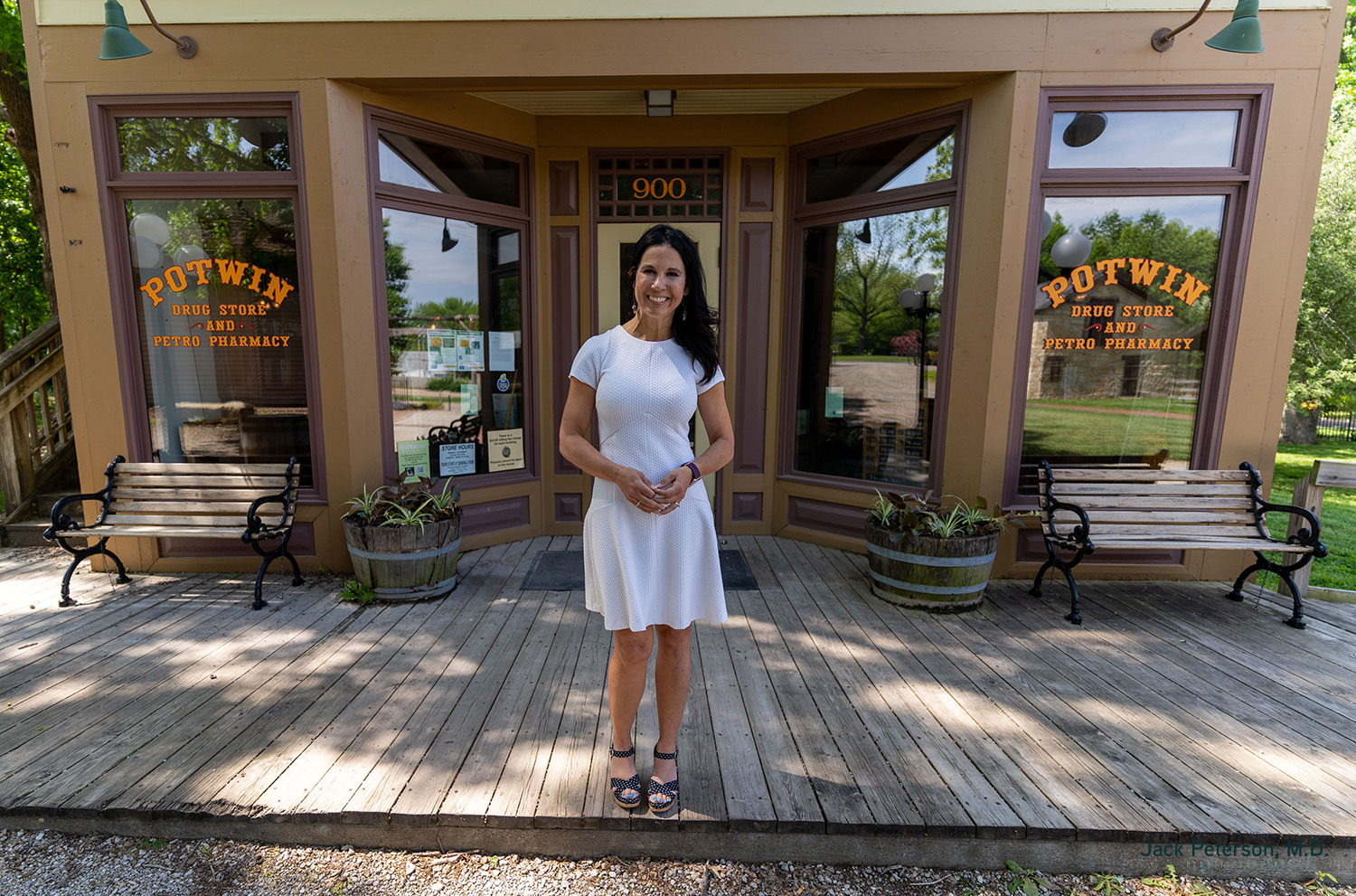 A woman in a white dress stands on a wooden porch in front of Potwin Drug Store and Petco Pharmacy, with benches and plants on either side—an inviting spot to discover personalized mommy makeover options.