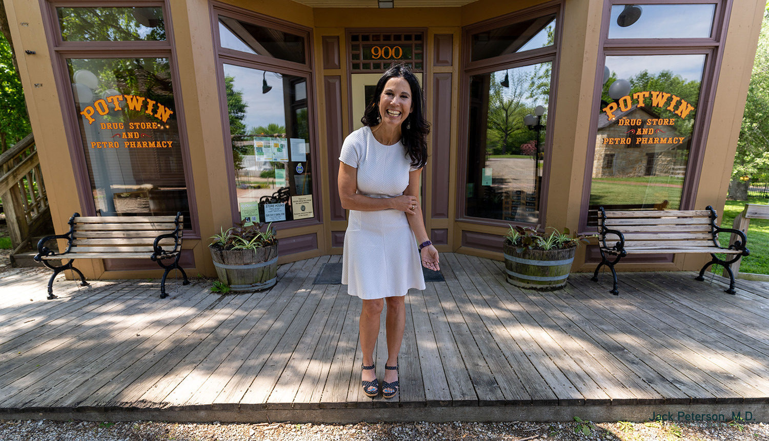 A woman in a white dress stands smiling on a wooden porch in front of Potwin Drug Store and Photo Pharmacy, known for offering skin treatments for acne scarring, with benches and potted plants on either side of the entrance.
