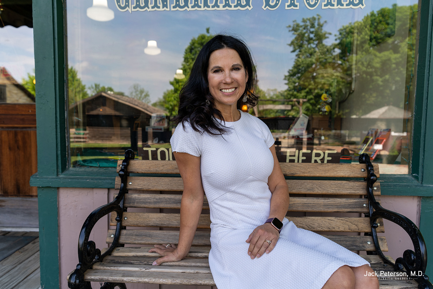 A woman in a white dress sits on a wooden bench outside, smiling, with her left arm resting on the bench and a smart watch on her wrist, exuding confidence that comes from exploring non-surgical facelift options.