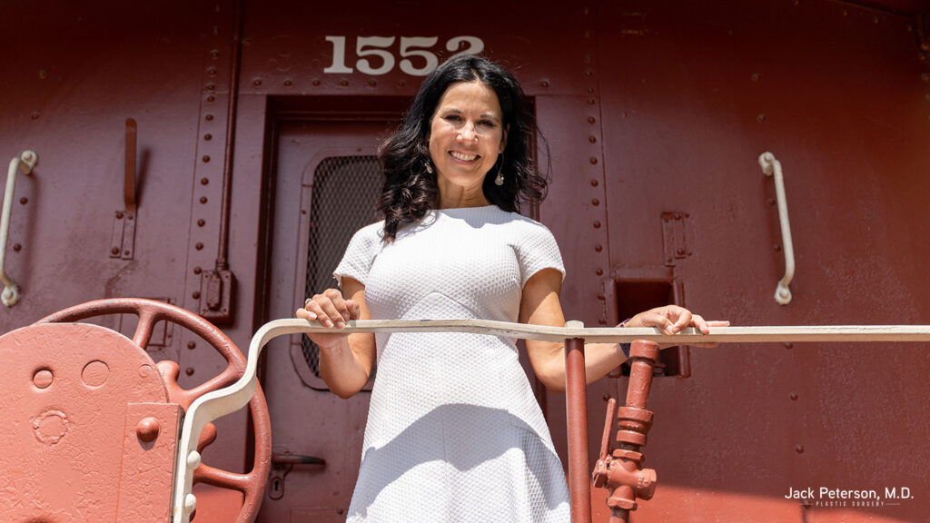 A woman in a white dress stands on the steps of red train car 1552, smiling at the camera—her confidence radiant, just like those exploring personalized mommy makeover options.