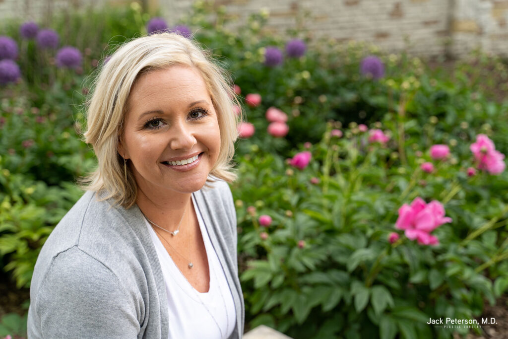 A woman with short blonde hair smiles while sitting in a garden with blooming pink and purple flowers, enjoying the confidence inspired by personalized mommy makeover options.