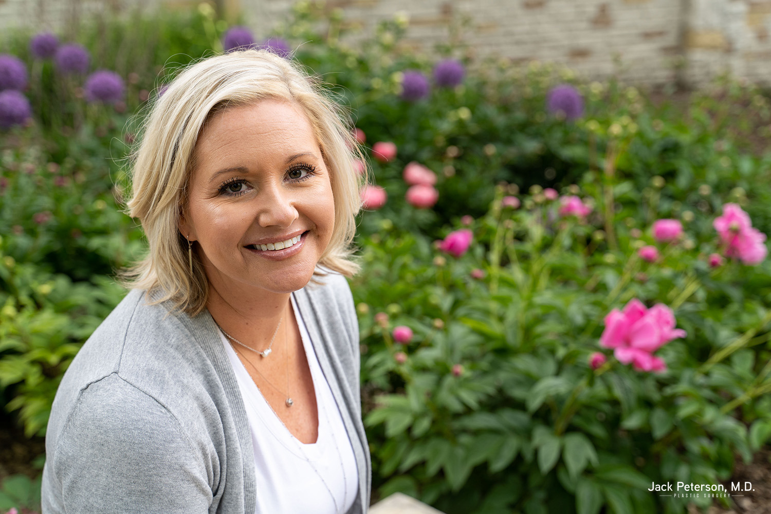 A woman with short blonde hair smiles while sitting in a garden with blooming pink and purple flowers, enjoying the confidence inspired by personalized mommy makeover options.