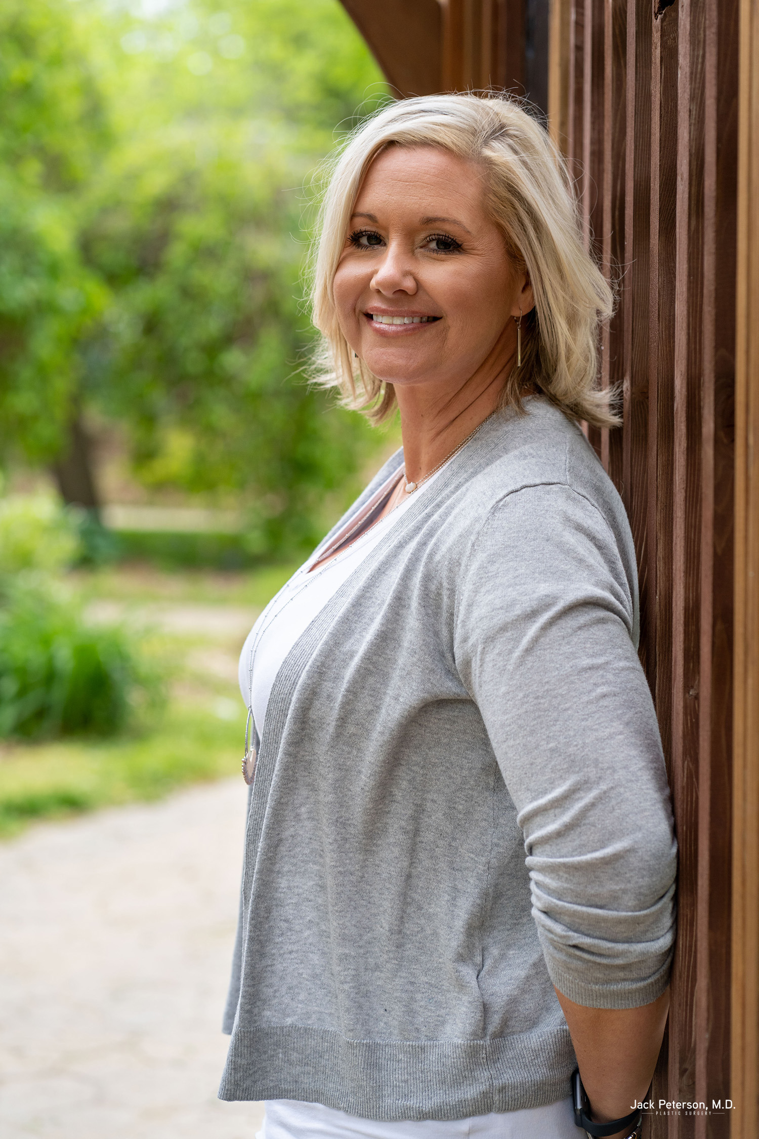 A woman with blonde hair, wearing a grey cardigan and white top, stands outdoors leaning against a wooden wall, smiling at the camera—showcasing confidence that comes from exploring personalized mommy makeover options. Greenery is visible in the background.