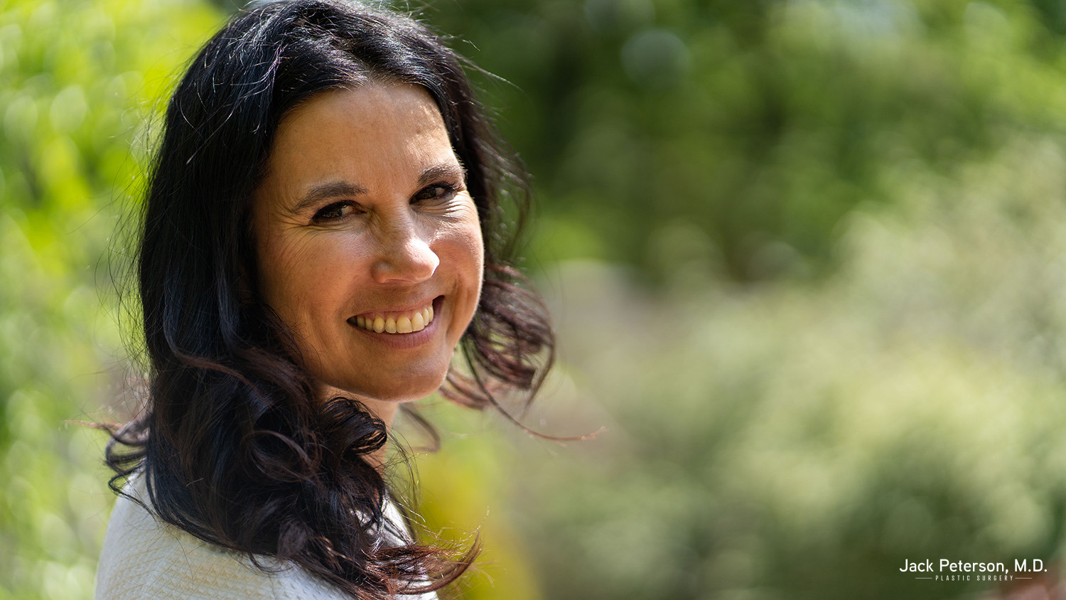 A woman with long dark hair smiles outdoors, wearing a light-colored top, radiating confidence after following her personalized mommy makeover plan, with a blurred green background.
