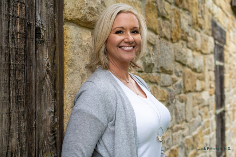 Woman with blonde hair wearing a gray cardigan and white top, smiling while leaning against a stone wall outdoors, embracing her confidence after choosing Motiva implants.