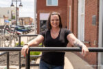Woman with short dark hair and tattoos stands smiling, leaning on a black railing outside on a sunny day in an urban setting, feeling confident after choosing the best breast implants for augmentation.