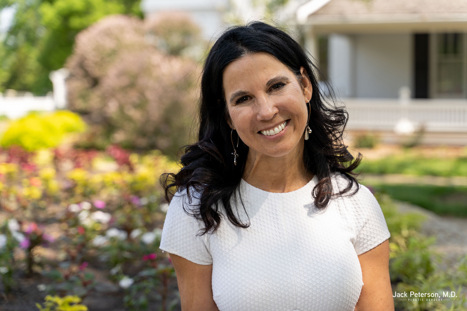 Woman with long dark hair wearing a white textured top smiles outdoors in a garden with flowers and a white building in the background, showcasing natural beauty enhanced by skin rejuvenation treatments. Text: "Jack Peterson, M.D." appears in the lower right corner.