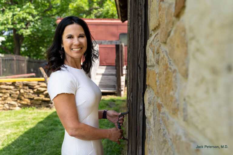 Woman in a white dress stands outside by a stone building, holding a wooden door handle and smiling confidently at the camera—much like what to expect from a tummy tuck: renewed self-assurance and poise.
