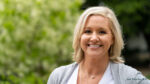 A woman with shoulder-length blonde hair smiles outdoors, wearing a gray sweater over a white top. Trees and greenery are blurred in the background, reflecting the confidence many feel after learning what to expect from a tummy tuck.