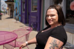 A woman with a tattoo on her arm, glowing with confidence, sits at a pink outdoor table on a sunny sidewalk. She smiles at the camera, colorful storefronts behind her—her look reflecting the results of the best breast implants for augmentation.