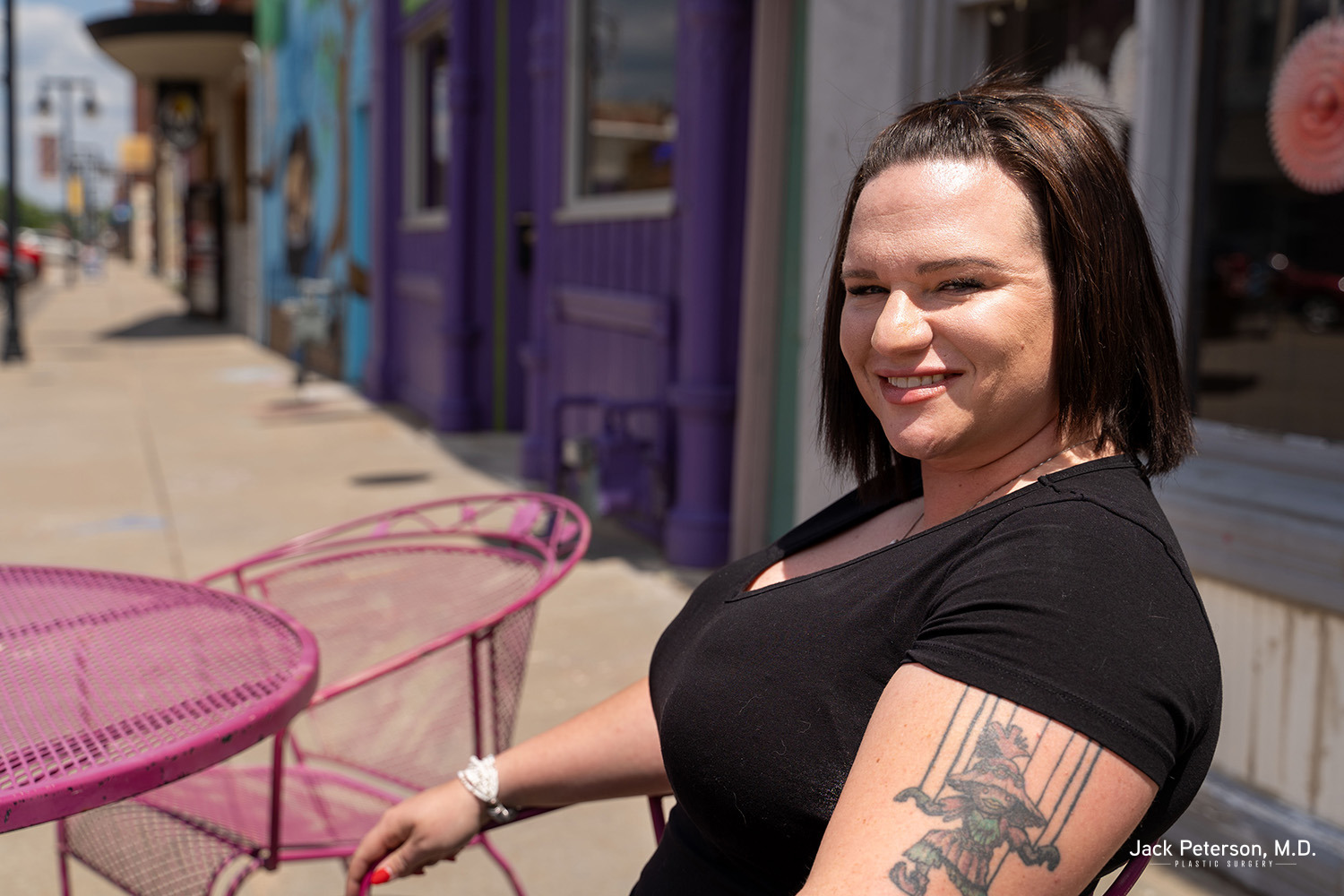 A woman with a tattoo on her arm, glowing with confidence, sits at a pink outdoor table on a sunny sidewalk. She smiles at the camera, colorful storefronts behind her—her look reflecting the results of the best breast implants for augmentation.